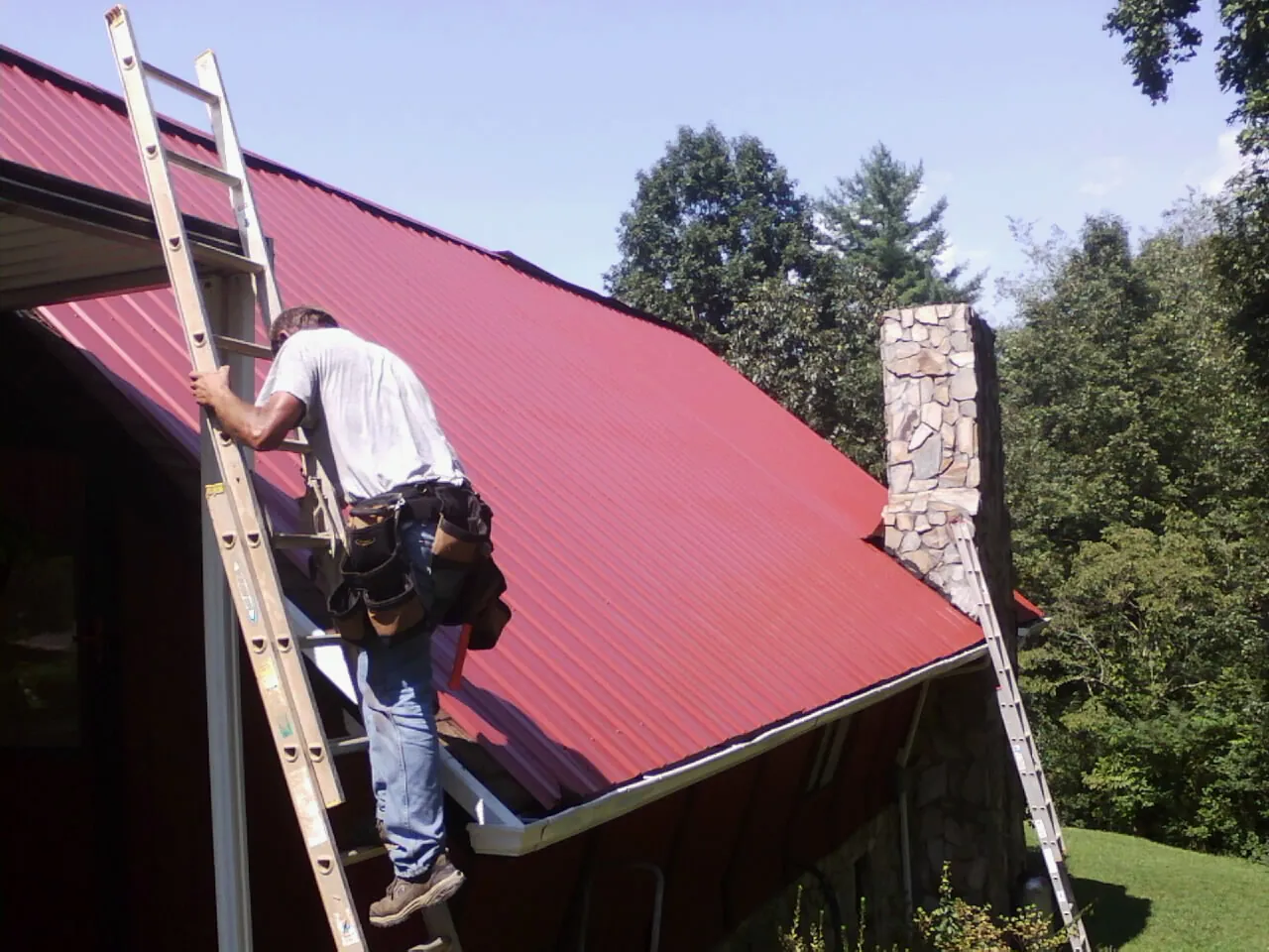 Roofer on a ladder beside a red metal roof and stone chimney — Soffit & Fascia Repair by Roofing Brothers in Marine